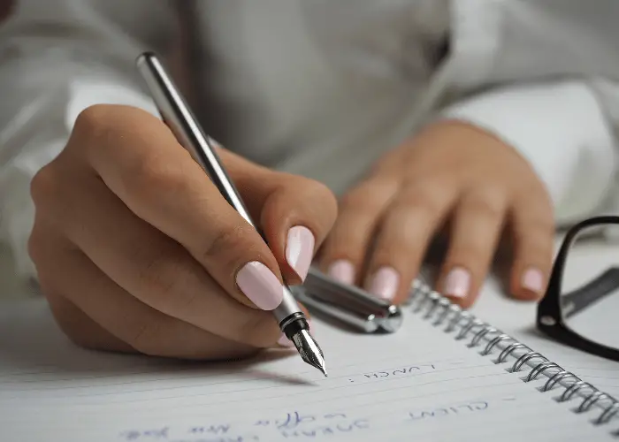 manos de una chica con uñas rosas escribiendo en un cuaderno con un estilógrafo