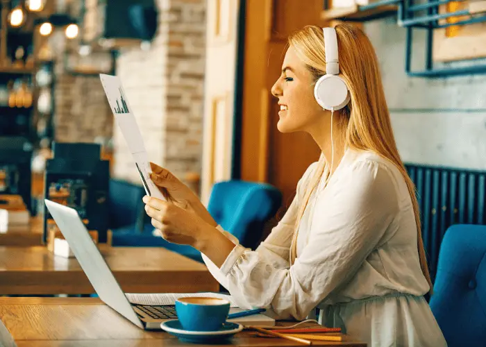 joven mujer estudiando / trabajando en una cafetería para ser más productiva, con cascos blancos y sujetando una hoja de papel con el portátil delante